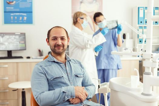 Portrait of man patient sitting on dental chair preparing for stomatology consultation waiting for somatology treatment. Senior woman doctor and medical nurse examining orthodontic x-ray