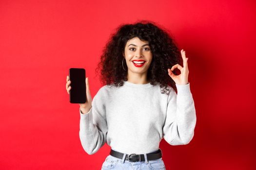 Cheerful european woman with curly hair, showing empty mobile phone screen and okay gesture, smiling satisfied, praise good app or promotion, red background.