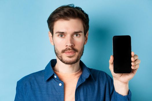 Close-up portrait of young man with beard showing empty smartphone screen with serious face, standing on blue background.