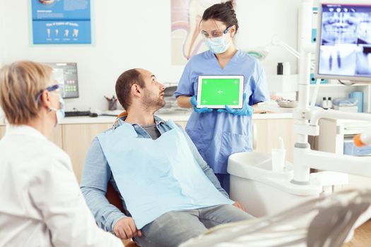 Sick man looking at mock up green screen chroma key tablet with isolated display while sitting at dental chair in hospital stomatology clinic office. Medical team explaining treatment toothache