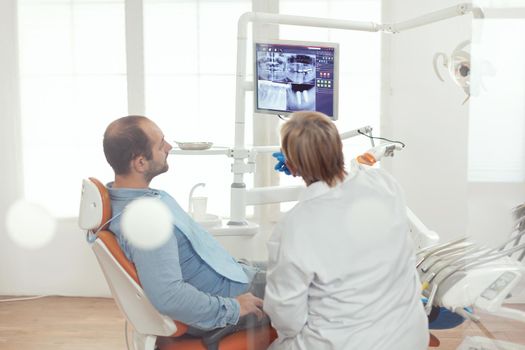 Stomatologist discussing with patient sitting in stomatological chair in dental clinic. Senior doctor explaining dental problem to sick man indicating x-ray digital image during dentistry appointment