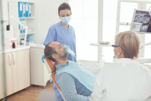 Hospital assistant putting oxigen mask before dental surgery sitting on stomatology chair in dentistry clinic . Senior orthodontist doctor and nurse working in modern office wearing mask and gloves