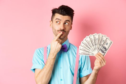 Man thinking about shopping, looking pensive while holding money in dollars, standing over pink background.