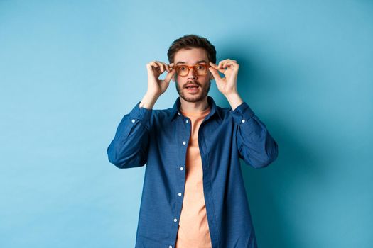 Image of handsome caucasian man put on glasses and looking at camera, trying new eyewear at optician store, blue background.