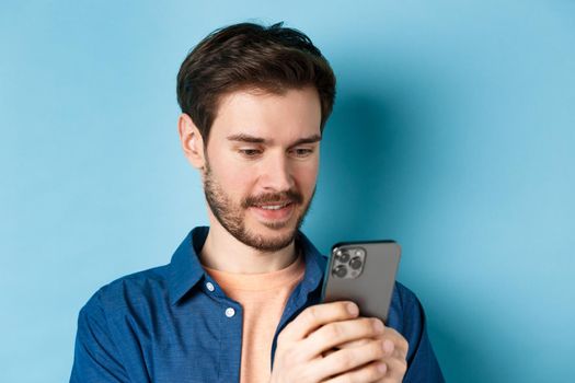 Close up portrait of happy young man reading mobile sreen, networking in smartphone, standing on blue background.