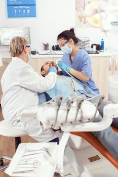 Stomatologist senior doctor doing toothache treatment on sick patient during stomatology consultation. Man sitting on orthodontic chair in hospital office while medical nurse cleaning teeh