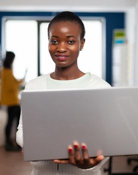 African videographer smiling at camera holding at laptop standing in creative agency office. Videographer working in multimedia studio production editing video in modern workplace.