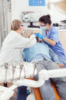 Orthodontist senior woman doing stomatology treatment on sick patient preparing for tooth surgery. Man siting on dental chair in hospital orthodontic office during medical procedure