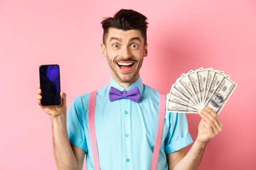 E-commerce and shopping concept. Cheerful guy in bow-tie showing empty smartphone screen and dollars money, smiling amazed at camera, standing on pink background.