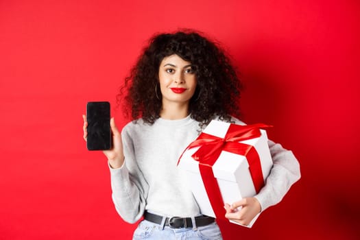 Beautiful woman with curly hair, showing shopping app on empty smartphone screen, holding gift wrapped in festive box, standing on red background.
