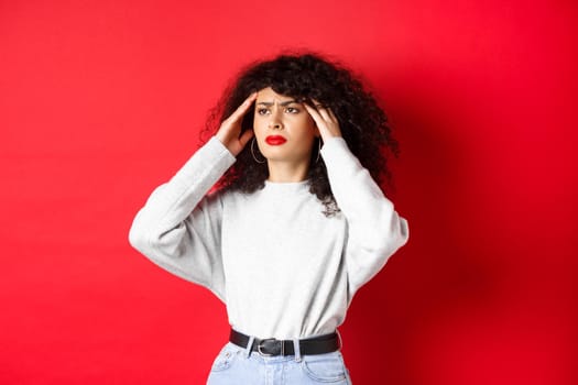 Distressed italian girl with curly hair, touching head temples and feeling bad, having headache, looking aside, standing on red background.