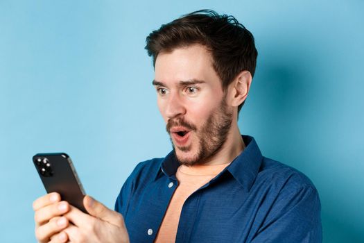 Image of happy young man stare at mobile screen, winning online and cheering, standing on blue background.