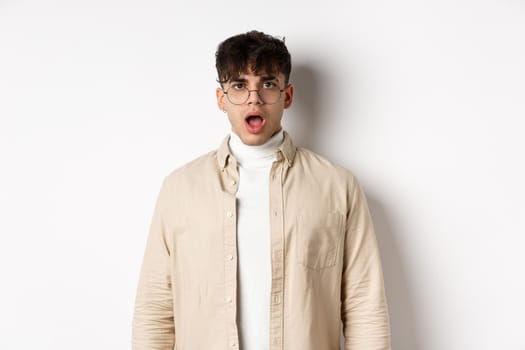 Portrait of shocked young man in glasses drop jaw, open mouth and stare amazed at camera, looking at something impressive, standing on white background.
