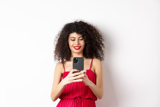 Happy fashionable woman taking photo on smartphone, photographing something on mobile and smiling, standing in red dress on white background.