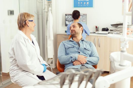 Senior dentist woman discussing with patient about toothache treatment during stomatological consultation in dental hospital office. Sick man sitting on dental chair waiting tooth radiograph