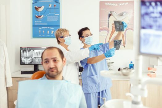 Sick man patient waiting for teeth examination sitting on dental chair in stomatology clinic room. Stomatologist doctor and medical nurse preparing for orthodontic surgery while examining radiography
