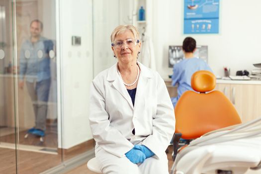 Orthodontist senior in medical uniform sitting on chair looking into camera waiting for man pacient to start stomatology treatment after tooth surgery. Team of dentists working in cabinet office