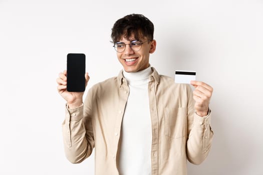 Online shopping. Handsome guy in glasses showing empty cellphone screen and credit card, smiling and looking at display pleased, standing on white background.