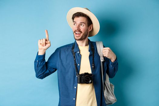 Handsome tourist looking and pointing up, travelling on summer vacation with backpack and camera, standing on blue background.