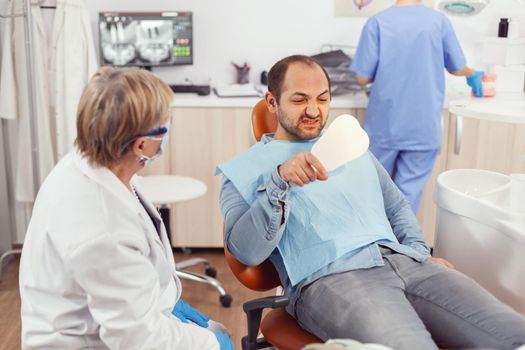 Sick patient inspecting teeth after dental surgery using mirror while sitting on stomatological chair in hospital clinic office. Senior stomatologist woman discussing about healdcare treatment