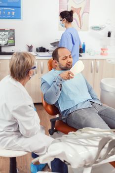 Senior stomatologist woman finishing dental surgery for man patient sitting on stomatology chair looking at tooth. Doctor explaining toothache treatment during stomatological consultation