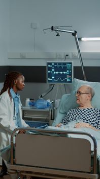 Caucasian nurse checking information files on patient while african american doctor talks to old man feeling sick in modern hospital ward. Multi ethnic medical staff healing disease