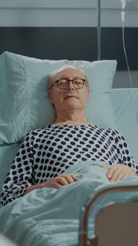 Hospital ward patient sleeping in bed at medical unit connected to nasal oxygen tube and rate monitor. Old man resting to heal pain after surgery procedure at intensive care facility