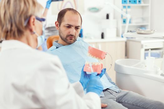 Stomatologist explaining proper dental hygiene using teeth skeleton during stomatology appointment. Dentist preparing sick man patient for dentistry surgery sitting on chair in examination room