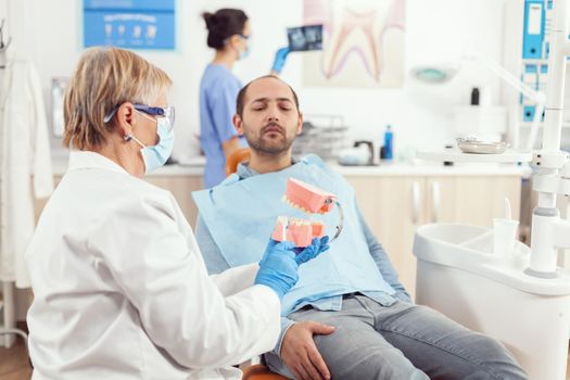 Senior stomatologist explaining to sick patient dental procedure using model of teeth. Doctor holding sample of jaw telling informations for healthy tooth while sitting in stomatology hospital clinic