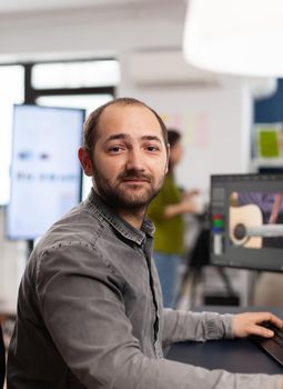 Man video editor looking at camera, working with footage and sound on computer with two displays. Videographer editing movie smiling while processing film montage sitting in startup office.