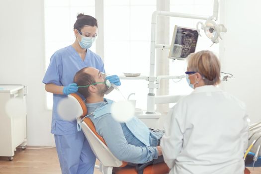 Medical assistant putting oxigen mask examining sick man patient for dentistry surgery while standing on dental chair in stomatology clinic room. Dentist doctor examining toohache