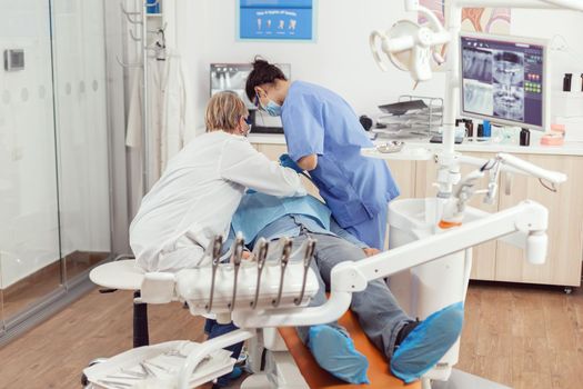 Stomatologist senior woman doing dental treatment on sick patient preparing denal tools for tooth surgery. Man siting on stomatology chair in hospital orthodontic office during medical procedure