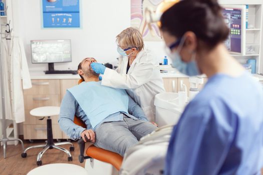 Sick man lying in stomatology chair with open mouth for medical exemination, in orthodontic clinic. Nurse preparing tools for oral problems working in stomatological hospital