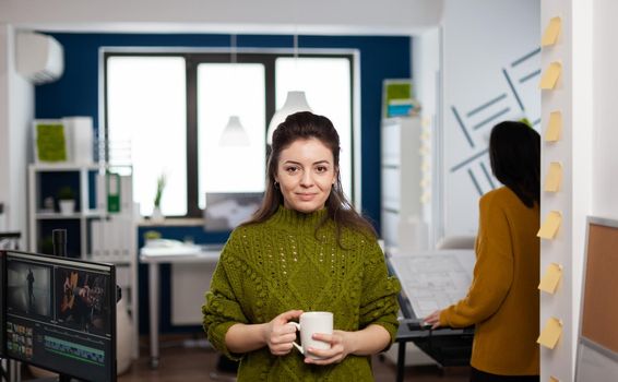 Portrait of creative designer smiling at camera holding a cup of coffee standing in start up agency office. Woman working in multimedia studio production editing video in workplace with diverse team.