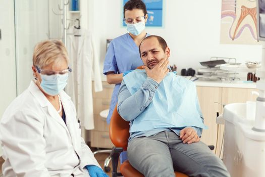 Man patient putting his hand on cheek showing toothache complaining about tooth pain. Senior woman explaining dental problem to doctor indicating mouth while sitting on stomatological chair