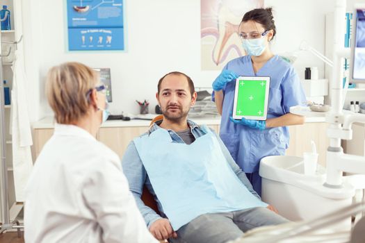 Stomatologist senior woman explaining tooth pain to patient while looking at mock up green screen chroma key tablet with isolated display. Medical team working in hospital dental office