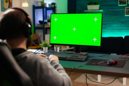 Professional cyber gamer playing digital videogames on powerful computer with green screen display. Player using pc with mock up chroma isolated desktop streaming shooter games wearing headset