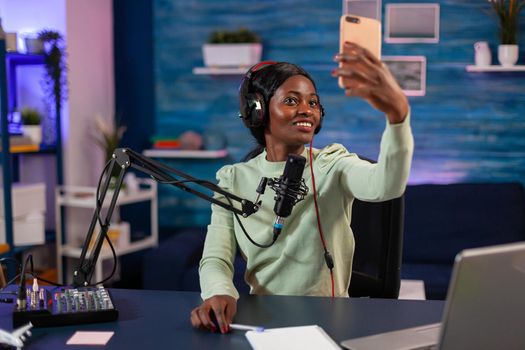 Afro woman taking selfie with smartphone and using professional gear to record episode in living room. On-air online production internet podcast show host streaming live content, recording digital social media.
