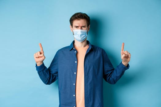 Covid and healthcare concept. Angry man in face mask pointing fingers up, feel disappointed and upset, standing on blue background.