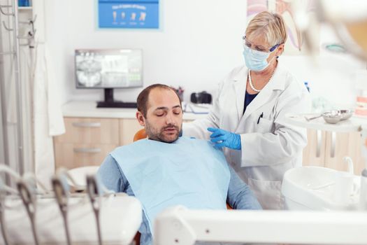 Senior stomatologist helping patient to stand up after dental surgery during medical stomatology clinic office. Orthodontist with protection face mask finising teeth inspection