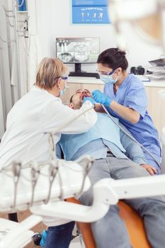 Senior woman stomatologist analyzing with mirror sick patient tooth health. Medical assistant preparing for cleaning teeth, treating affected mass while sick man sitting on stomatology chair
