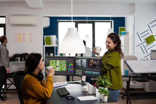 Happy coworkers talking about film montage looking at movie footage working in creative start up agency office with two monitors. Videographers processing video project in post production software