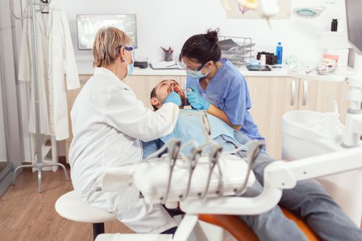 Sick patient sitting in stomatology chair with open mouth for inspection, in stomatological office, doctor preparing for oral surgery. Dentist and medical nurse with masks removes tooth