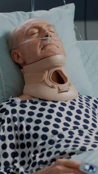Portrait of elder patient sitting with cervical neck collar in hospital ward bed at intensive care medical clinic. Old man with nasal oxygen tube and IV drip bag resting after surgery