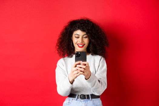 Smiling modern girl taking photos on smartphone, looking at screen and recording video, standing against red background.