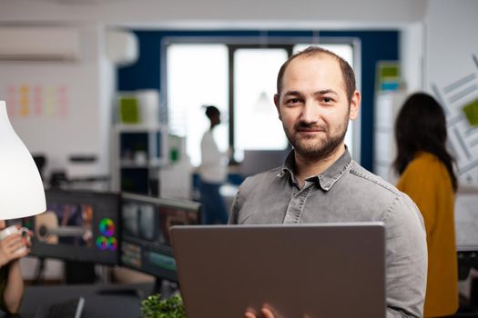 Video editor worker standing in front of camera smiling working in creative agency office holding laptop. Man videographer works in multimedia studio production editing video in modern workplace.