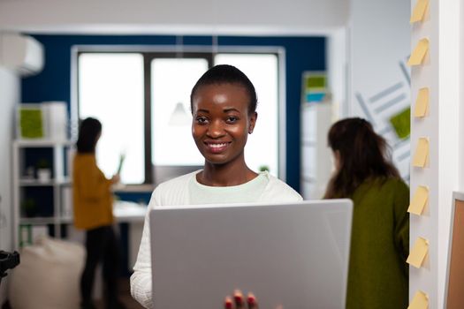 Close up of african woman looking at camera smiling standing in start up creative agency office holding laptop, typing on it. Black videographer editing video project using post production software