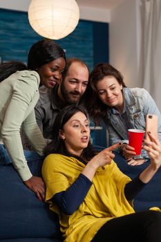 Multiracial friends hanging out late at night watching funny entertainment video on smartphone. Group of multiracial people spending time together sitting on couch late at night in living room.