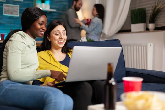 Mixed race friends talking while looking at funny video series on laptop sitting on couch in living room. In background woman and man drinking beer enjoying time together during fun party.