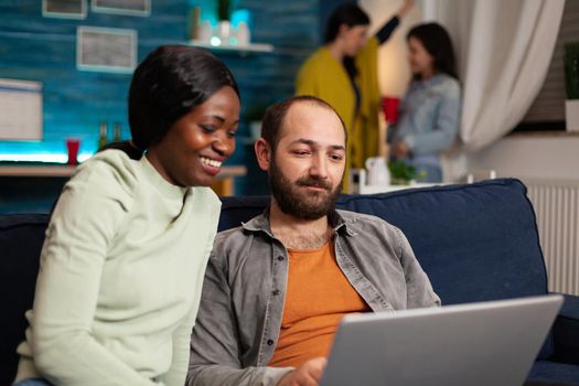 Multiracial friends socializing while watching online videos on laptop relaxing on sofa. In background two women drinking beer enjoying time spend together during entertainment party.